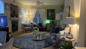 A living room with a fireplace, a television, a ceiling fan, and a round coffee table with decorative items. - Lovely 2 bedroom craftsman style home (Lincoln Beach)