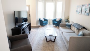Una sala de estar con una televisión de pantalla plana, una mesa de centro, dos sillones azules, un sofá color beige y un ventilador de techo.