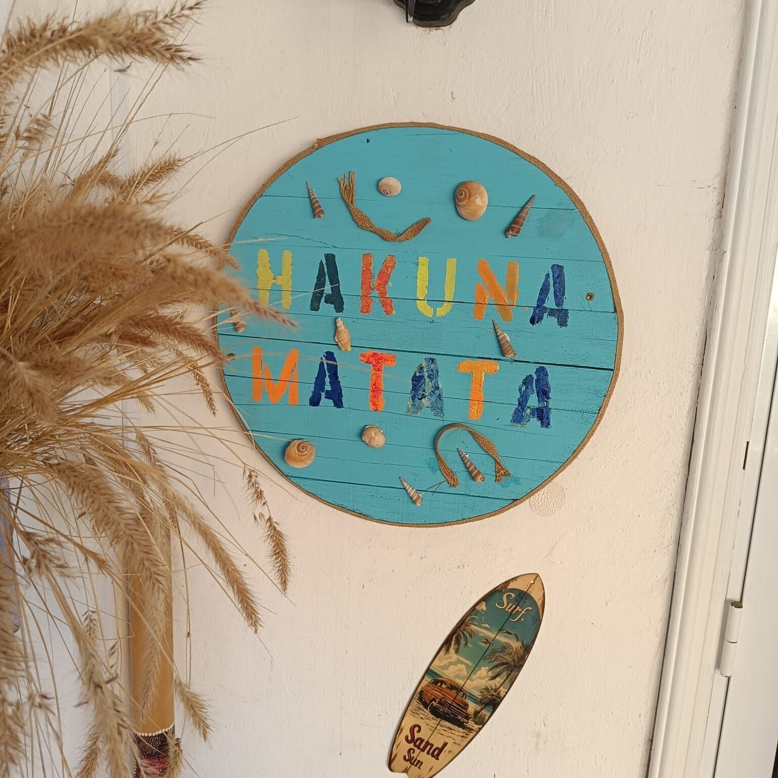 A round sign with the text "HAKUNA MATATA" surrounded by seashells and a surfboard.