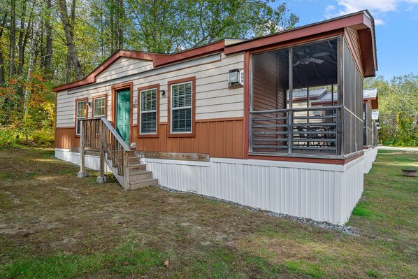 A single-story mobile home with a front porch, a green door, and a large windowed porch area. - Cozy Cottage at Lake George Campsites! (Queensbury)