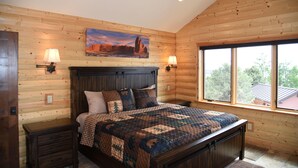 Ein Schlafzimmer mit Holzwand aus Baumstämmen, einem großen Bett mit Decke, zwei Nachttischlampen und einem Fenster mit Blick auf Bäume.