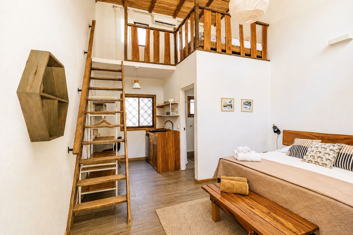 A bedroom with a wooden bed, a bench, a ladder, and a view of the kitchen and bathroom.