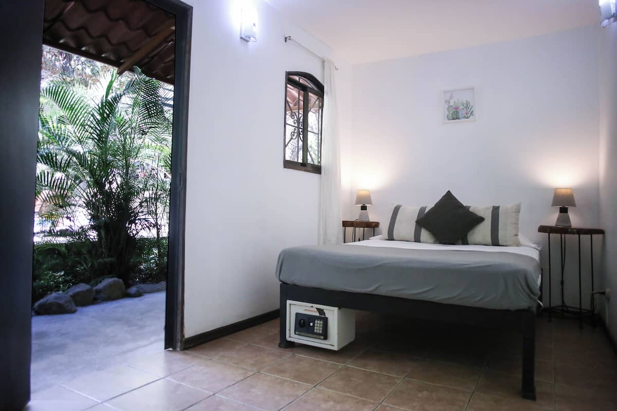 Une chambre à coucher avec un lit, deux lampes de chevet, une fenêtre avec des rideaux, un petit tableau accroché au mur et une vue sur la verdure à l’extérieur.