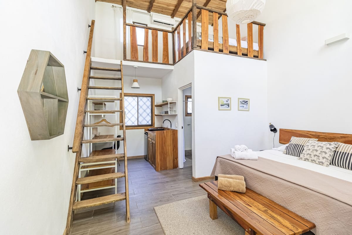 Une chambre avec un lit en bois, un banc en bois, une échelle en bois et, au fond, un coin cuisine.