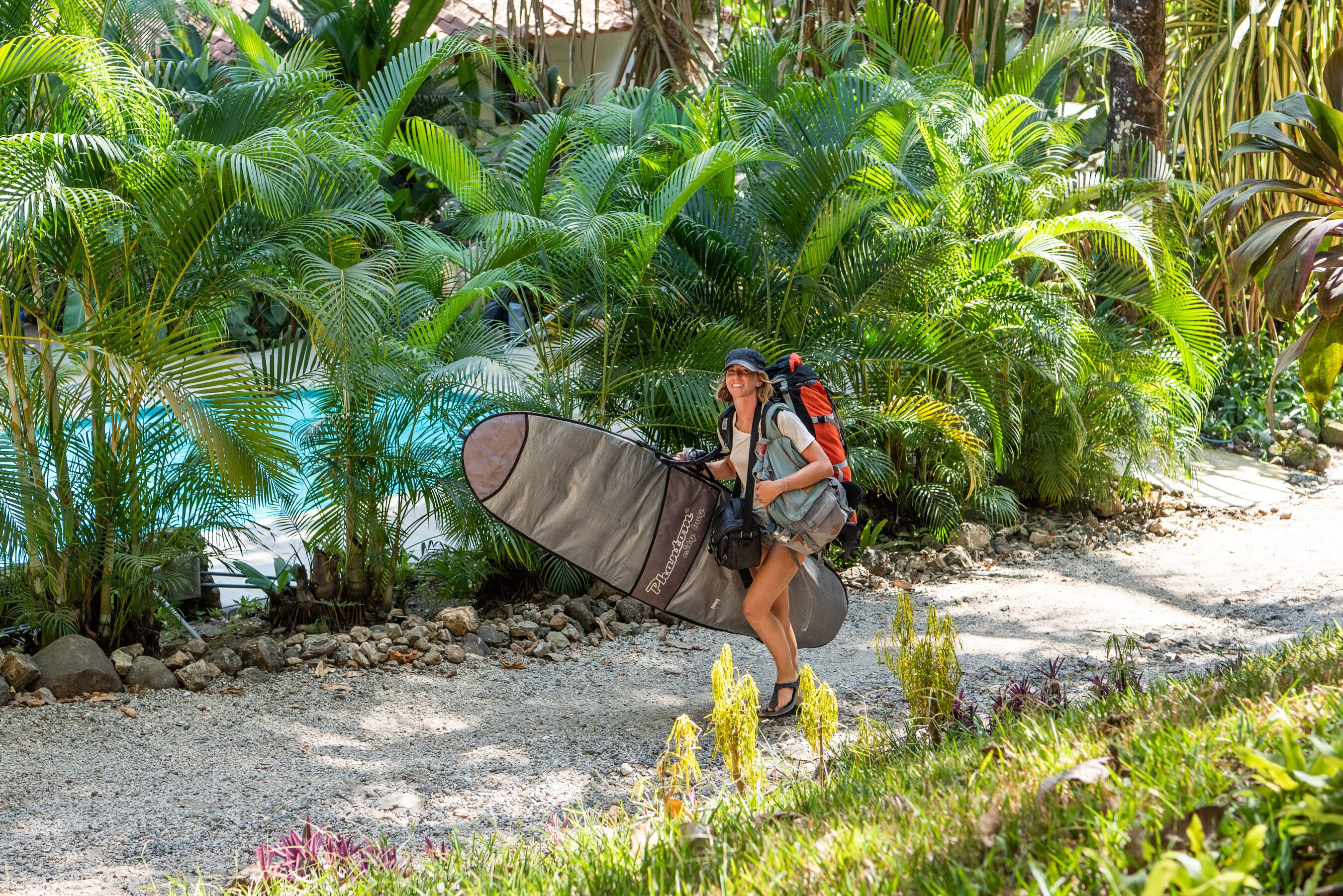 A person carrying a large surfboard and a backpack, walking on a gravel path surrounded by lush greenery.