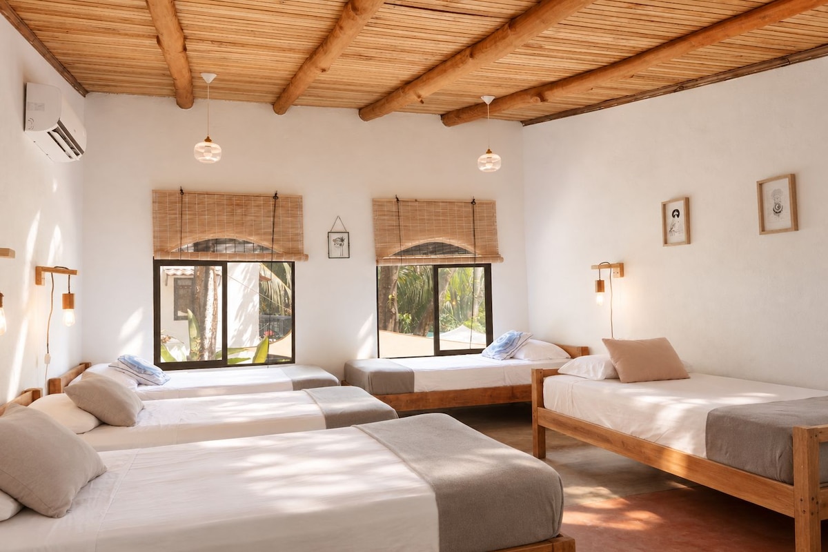 A room with three wooden beds, a ceiling with exposed beams, and large windows with blinds.