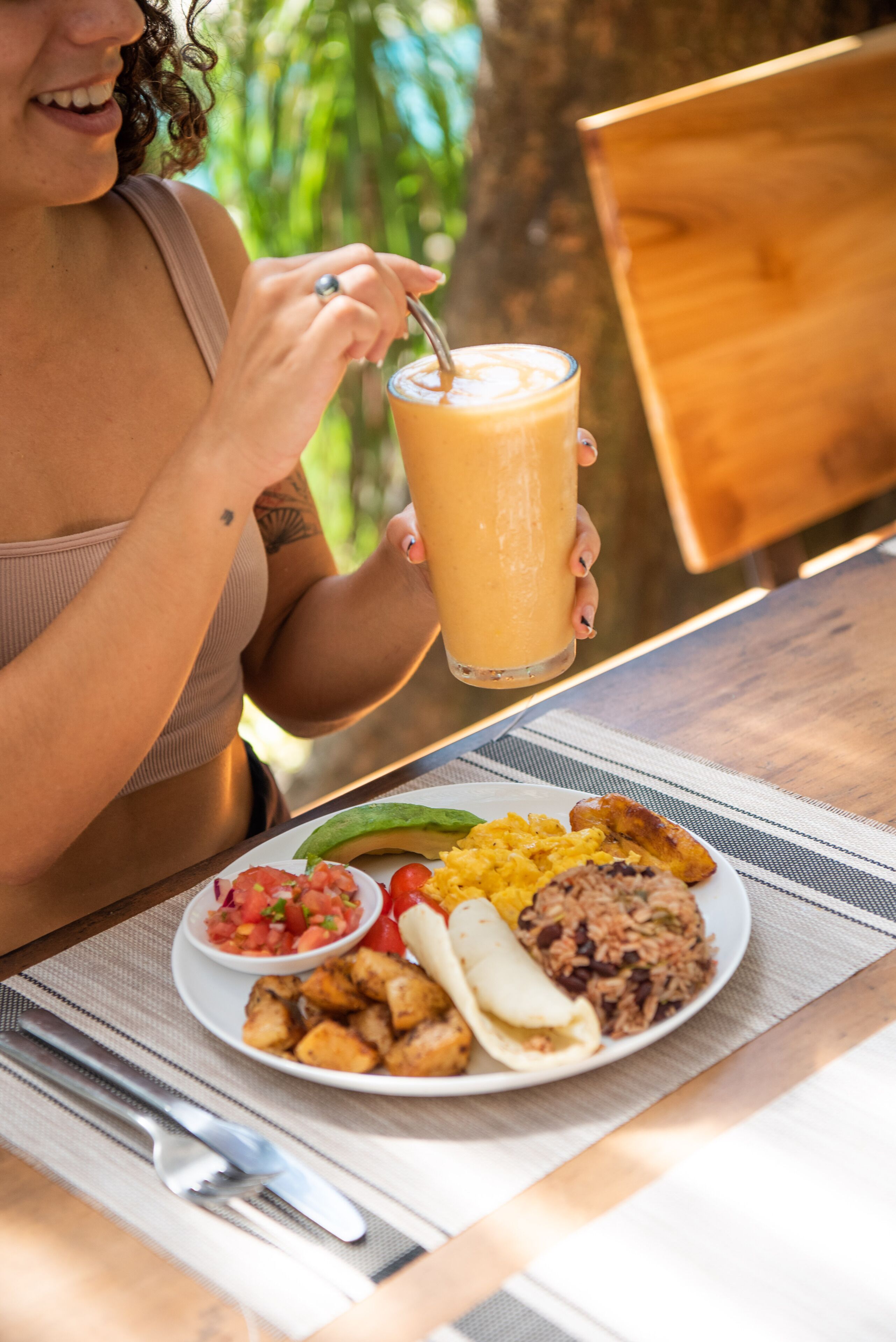 Una persona sosteniendo un vaso de batido, sentada a una mesa con una comida que incluye arroz, frijoles y verduras.