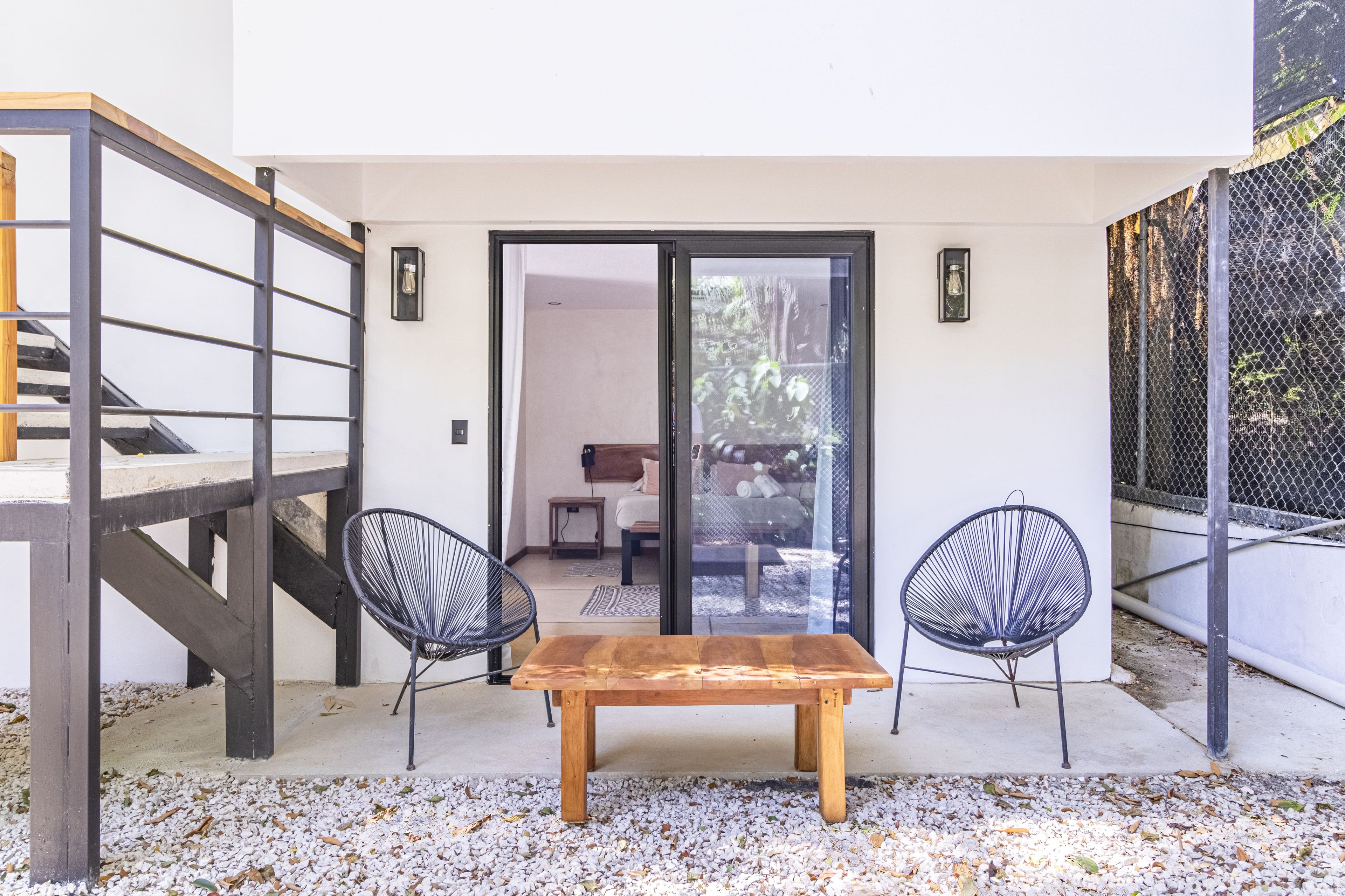 A patio with a wooden table and two chairs, a staircase, and a view into a room with a bed and a window.