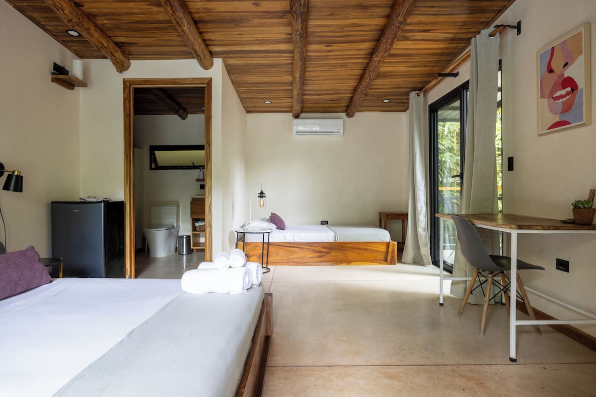 Une chambre d’hôtel avec deux lits, un bureau, une chaise et une petite table. Elle est équipée d’un réfrigérateur, d’un lavabo et d’une fenêtre avec des rideaux.