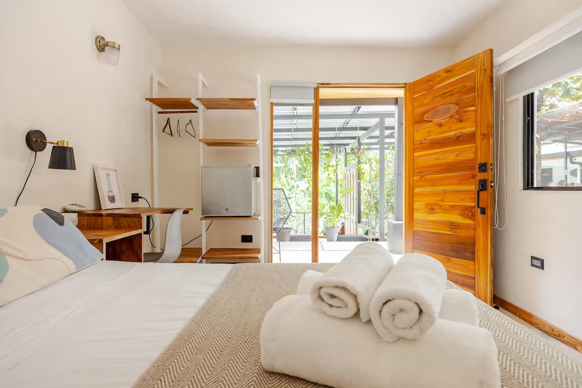 A modern bedroom with a bed, a desk, a chair, a TV, and a wooden door leading to a balcony.