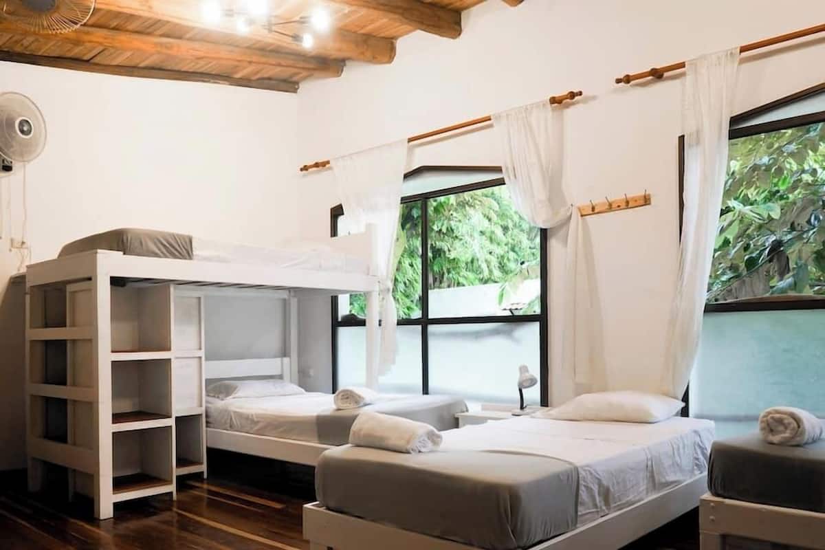 Une chambre avec des lits superposés, un ventilateur, une fenêtre avec des rideaux et une vue sur la verdure.