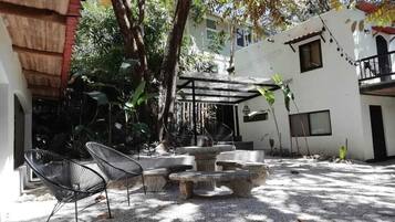 Un área de estar al aire libre con bancos de piedra y sillas de metal, rodeada de árboles y un edificio blanco con balcones.