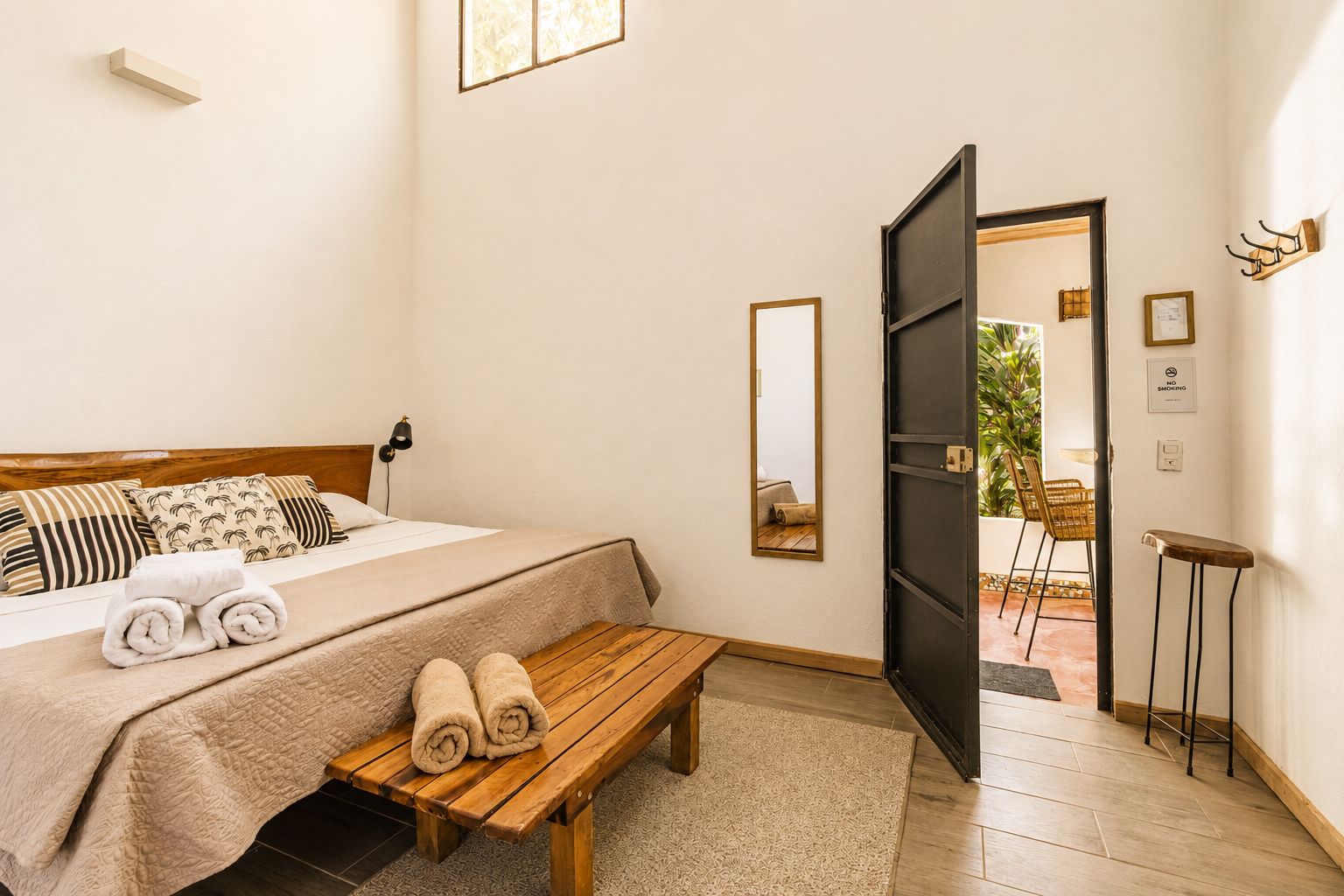 A hotel room with a bed, a bench, a mirror, and a door leading to an outdoor area with chairs and plants.