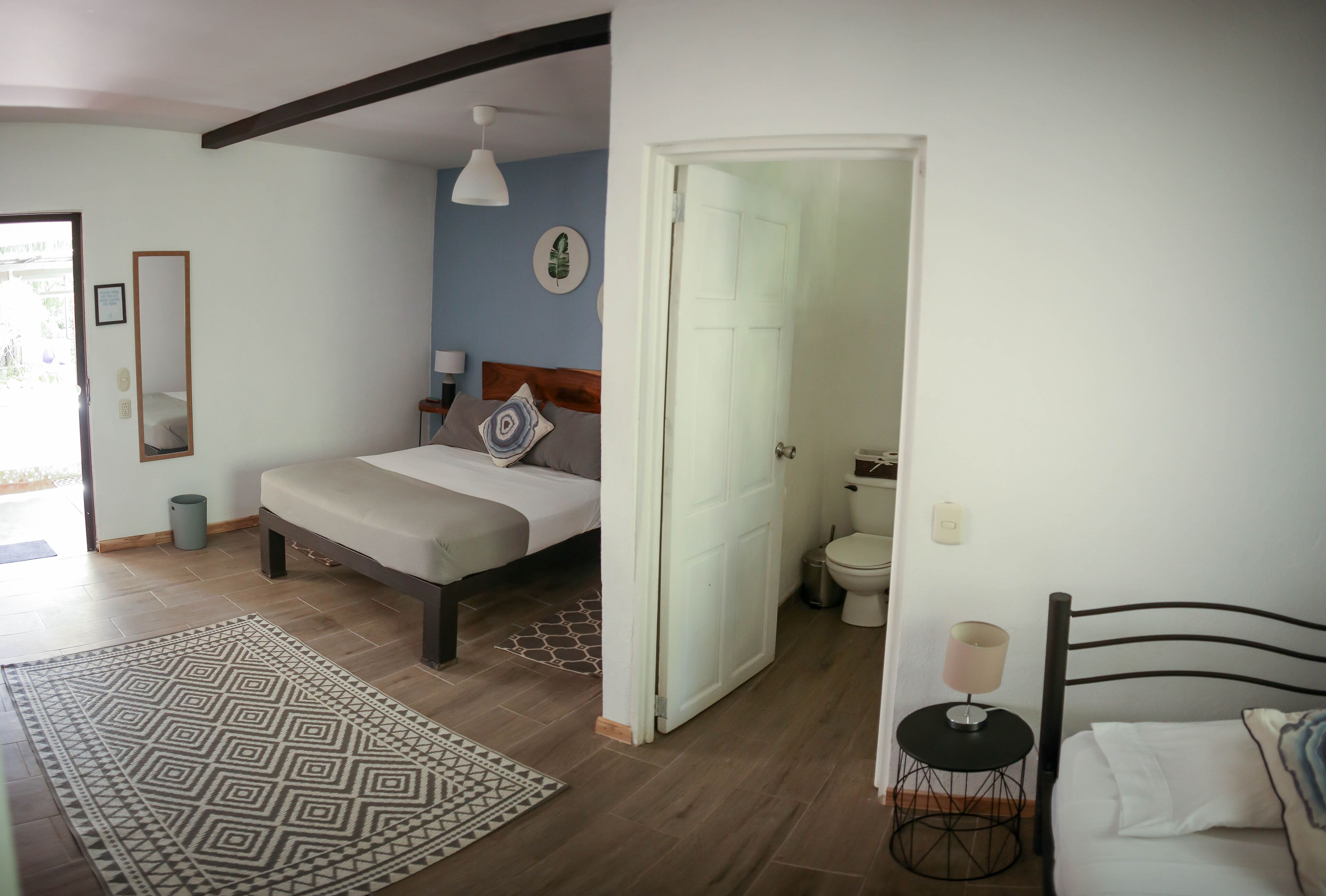 A bedroom with a bed, bedside table, and a nightstand. There is a bathroom with a toilet and sink. A door leads to another room.
