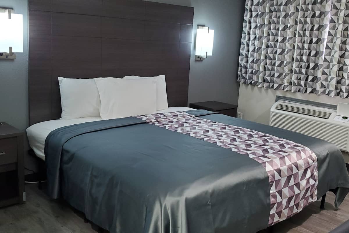 A hotel room with a bed, a nightstand, a wall-mounted air conditioner, and patterned curtains.