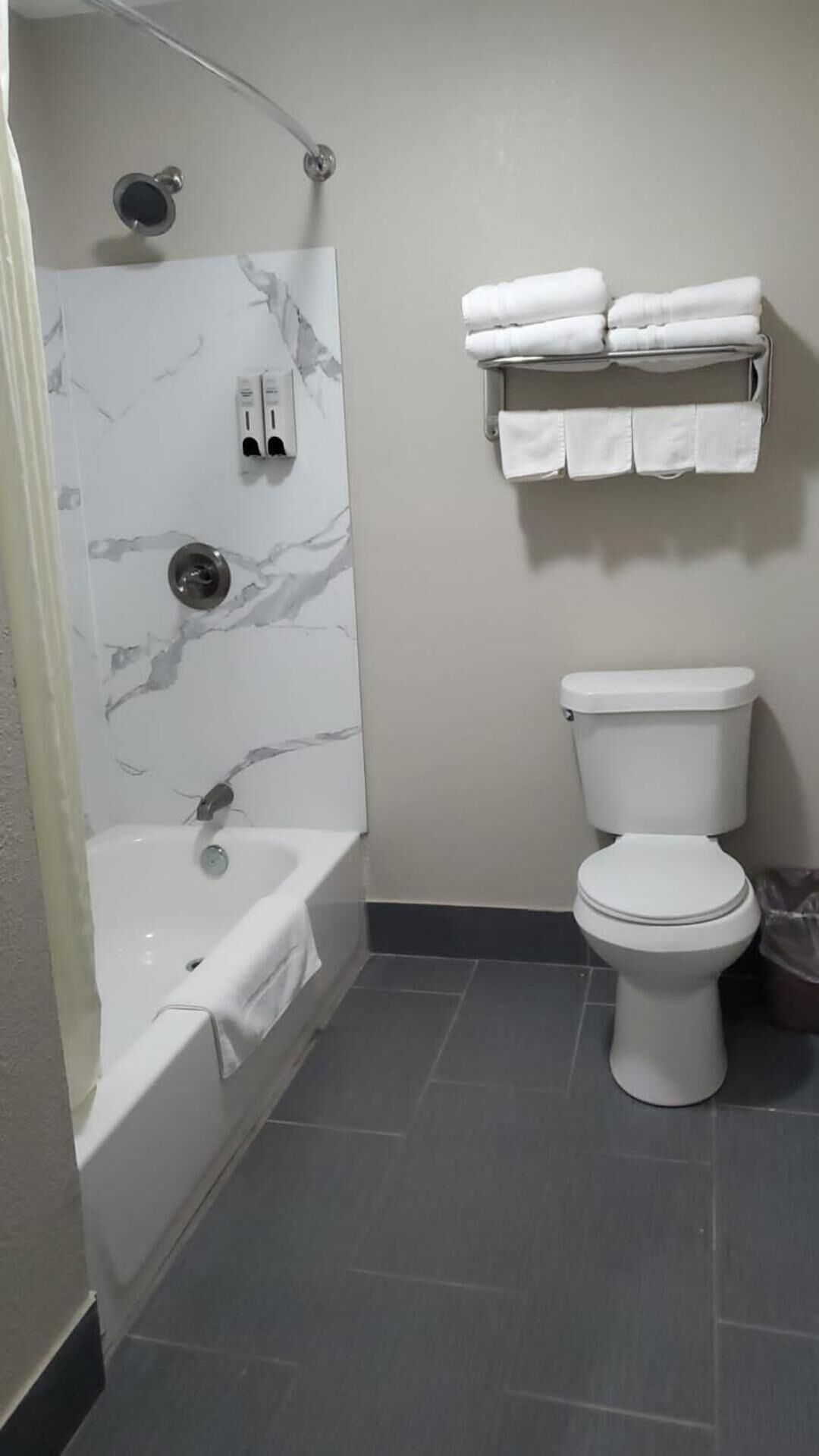 A bathroom with a marble shower wall, a bathtub, a toilet, and a towel rack with folded towels.