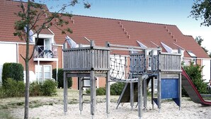 Een houten speelplaats met een glijbaan en klimconstructies, gelegen voor een woongebouw met een rode dakbedekking.
