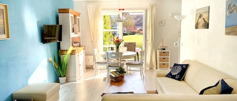 A living room with a sofa, a TV, a dining table with chairs, and a view of the outdoors.