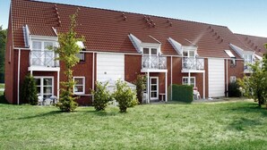 Een woongebouw met twee verdiepingen, voorzien van een rode pannendak, ramen met witte kozijnen en een kleine voortuin met gazon en enkele bomen.