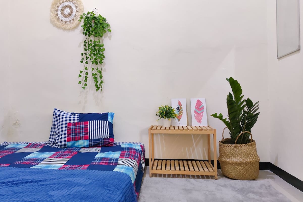 Ein Schlafzimmer mit einer blauen gesteppten Bettdecke, einem karierten Kopfkissen, einem kleinen Holfnachtkästchen mit einer Pflanze und einem gewebten Korb.