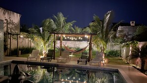 Une piscine entourée de chaises longues et d’un hamac suspendu sous des guirlandes lumineuses, le tout bordé de palmiers et d’un jardin.