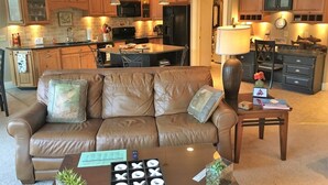 A living room with a brown leather sofa, a coffee table with a remote control, and a lamp. - This riverfront downtown condo boasts and open floor plan for kitchen, living room, and dining areas. Or step onto the balcony over the Arkansas River to hear the water rushing by. Oversized master suite has its own balcony and indoor sitting area. (Salida)