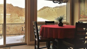 A dining area with a table covered in a red tablecloth, chairs, a chandelier, and a view of a desert landscape through large windows. - This riverfront downtown condo boasts and open floor plan for kitchen, living room, and dining areas. Or step onto the balcony over the Arkansas River to hear the water rushing by. Oversized master suite has its own balcony and indoor sitting area. (Salida)