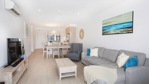A modern living room with a grey sofa, a coffee table, and a TV on a wooden cabinet. There is a dining area with a white table and chairs, and a kitchen in the background.