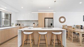 A modern kitchen with a central island, wooden stools, a dining area with a wooden table and chairs, and a stainless steel refrigerator.