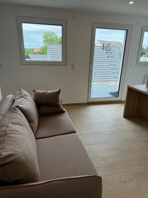 A modern living room with a large beige sofa, two windows with a view of a residential area, and a wooden floor.