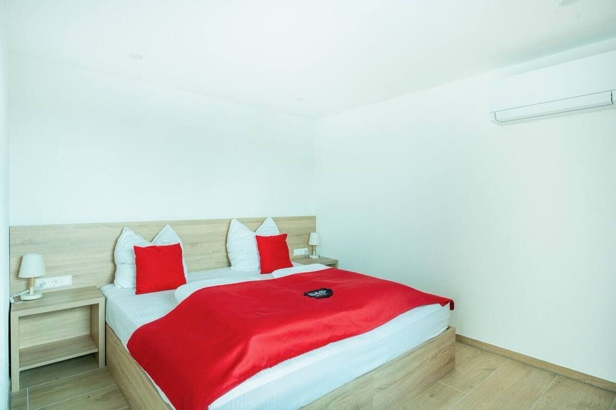 A hotel room with a wooden headboard, a bed with white and red bedding, a red throw blanket, a nightstand with a lamp, and a wall-mounted air conditioning unit.