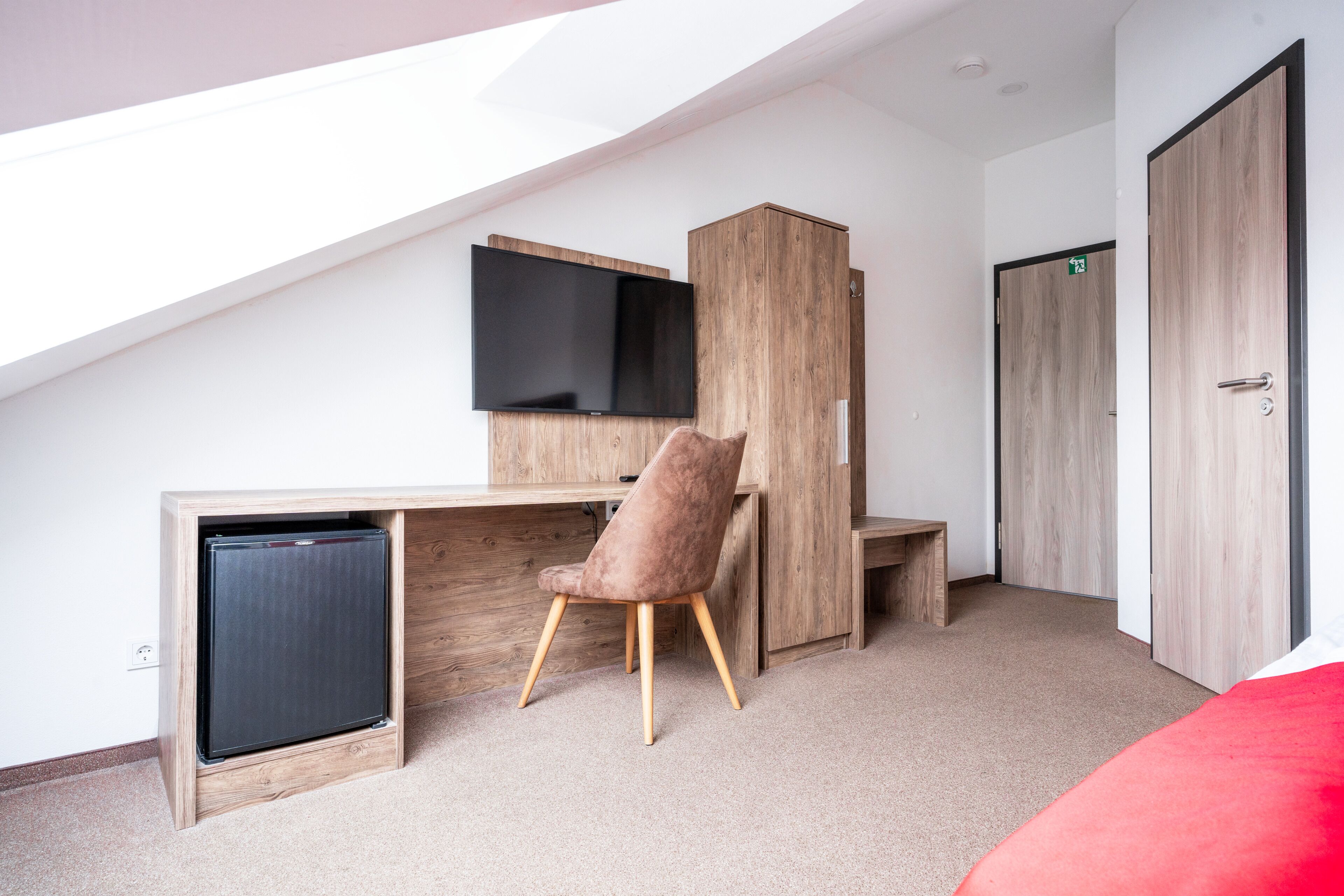 A modern room with a sloped ceiling, a wooden desk with a flat-screen TV, a cushioned chair, a small built-in fridge, and a wardrobe.