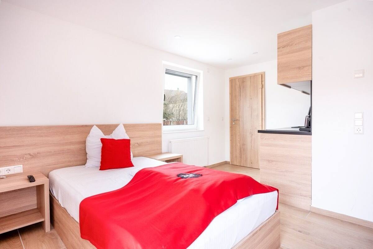A modern hotel room with a wooden headboard, a bed with white and red bedding, a red throw blanket, a nightstand, a small table, and a window providing a view of the outdoors.