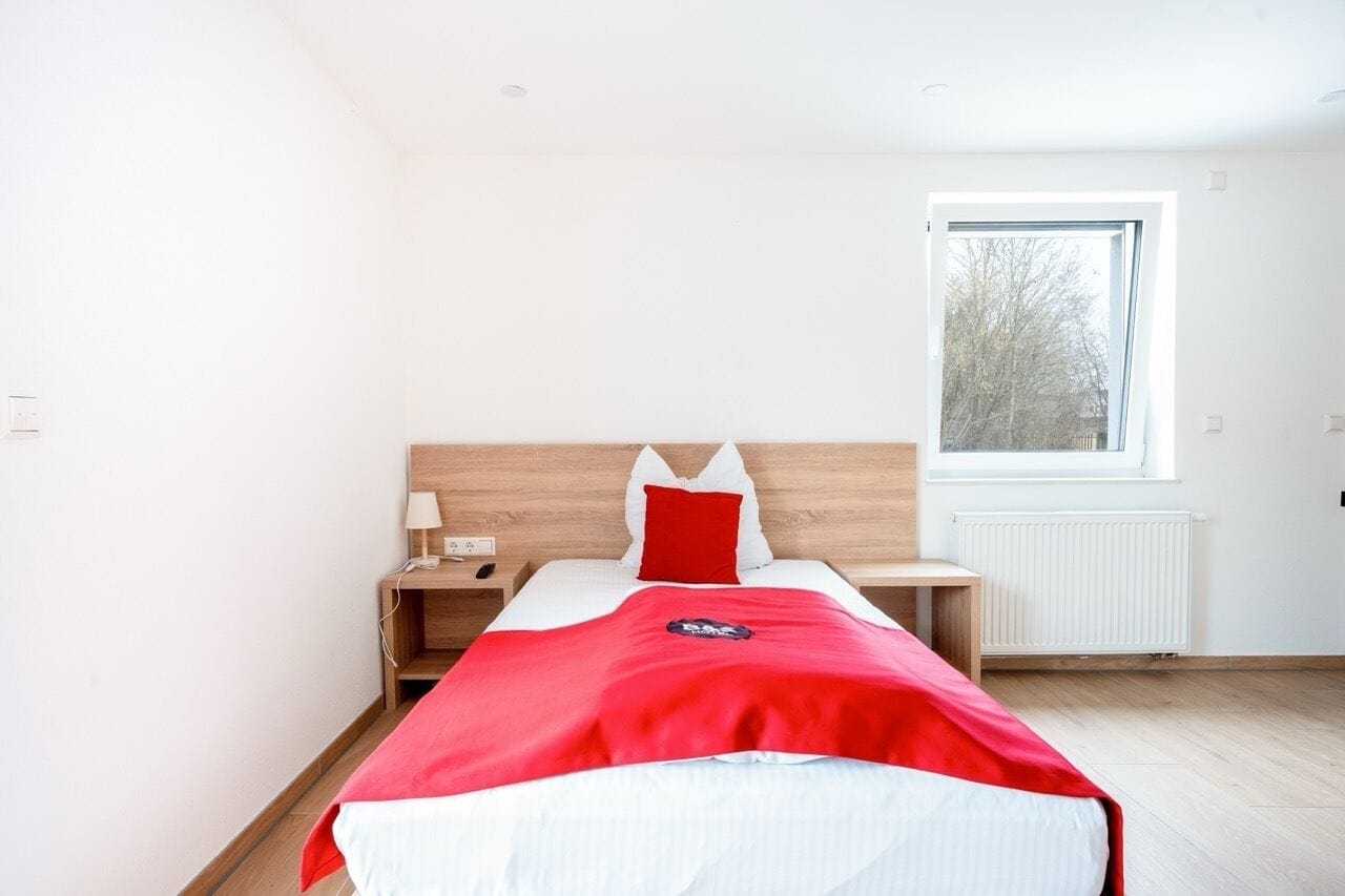 Een moderne hotelkamer met een houten hoofdbord, een groot raam, een nachtkastje met een lamp en een rode sprei.