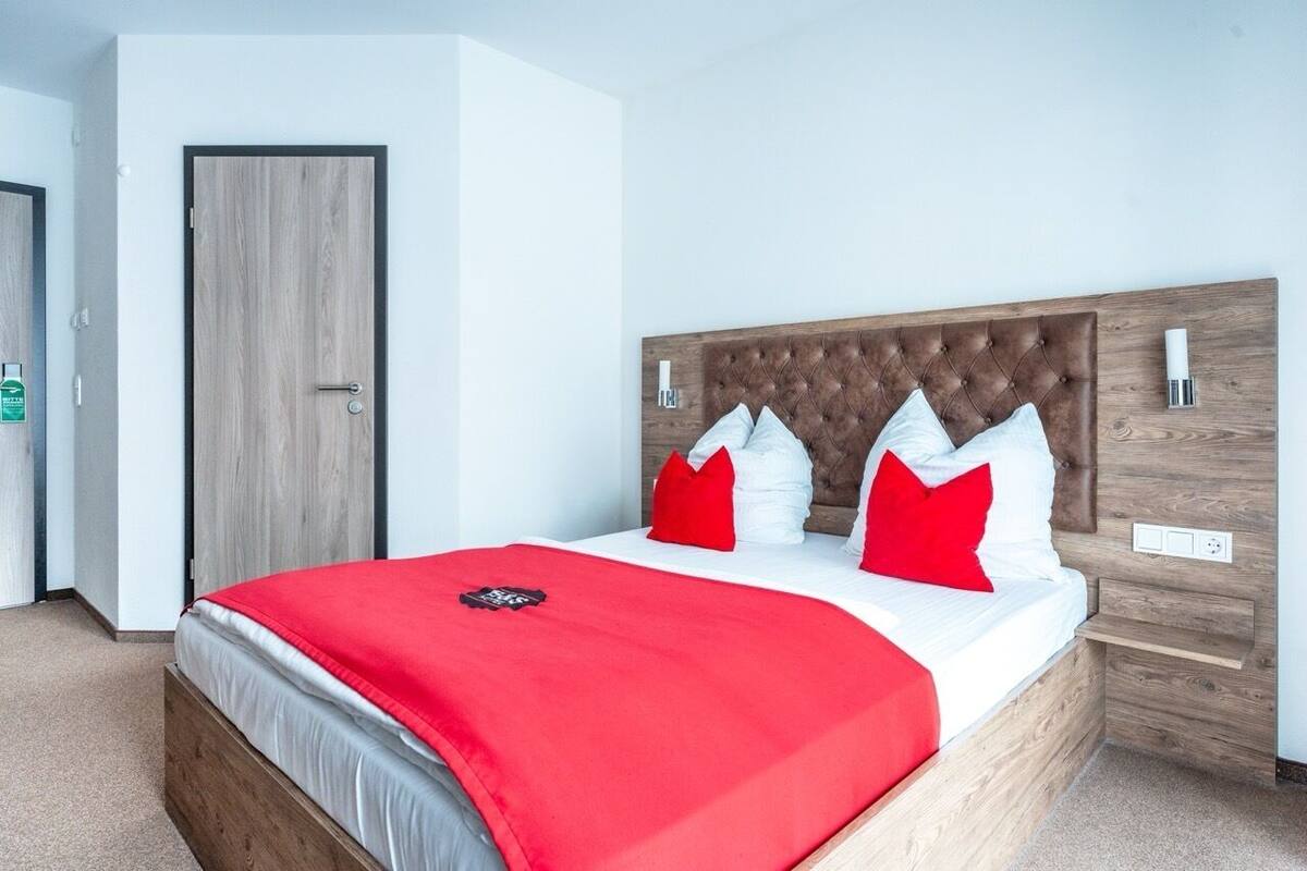 A bedroom with a wooden headboard, a bed with red and white bedding, a red throw blanket, and a nightstand with a lamp.