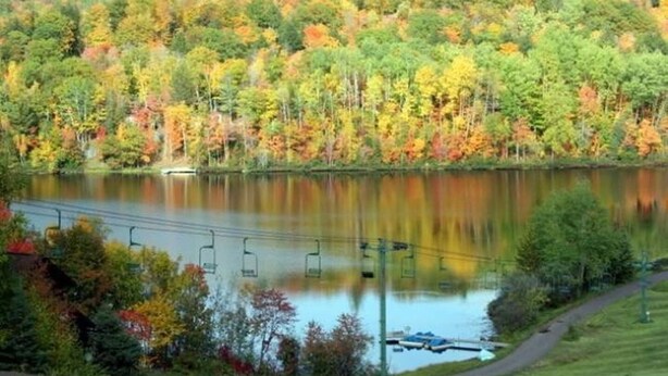 Ein See, umgeben von Bäumen mit Herbstlaub, eine Anlegestelle und eine Sesselbahn.