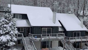 Eine zweistöckige Lodge mit schneebedecktem Dach und Holzfassade. Zwei auffällige Treppen führen zum Eingang, und ein kleiner Vorbereich bietet eine Sitzbank.