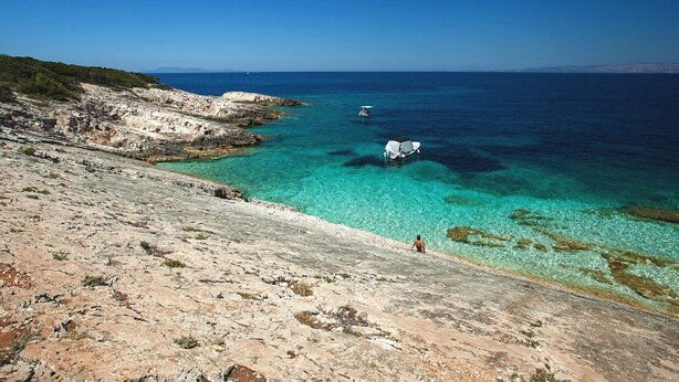 Une côte rocheuse bordée d’eaux turquoise et limpides, un bateau et une personne qui se promène le long de la plage.