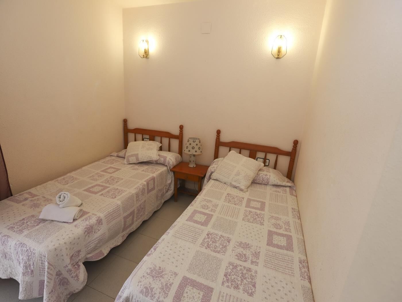 Habitació petita i senzilla amb dos llits individuals, cadascun amb un capçal de fusta, una tauleta de nit amb una làmpada i dues làmpades murals.