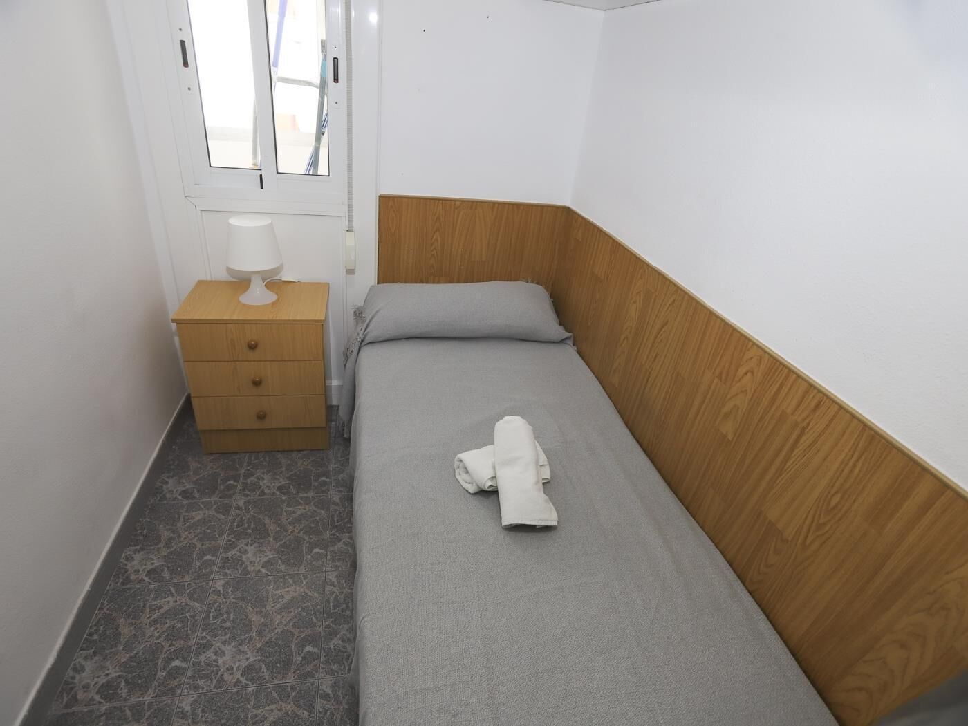 A small, single-bed room with a wooden headboard, a bedside table with a lamp, and a window.
