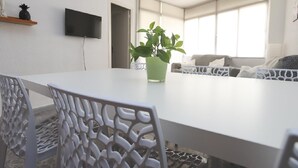 Ein modernes Wohnzimmer mit einem weißen Tisch, weißen Stühlen, einer Zimmerpflanze, einem Flachbildfernseher und einem Fenster mit Jalousien.