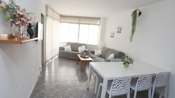Ein modernes Wohnzimmer mit einem grauen Sofa, weißen Sesseln, einem Esstisch und einem großen Fenster, das für natürliches Licht sorgt.