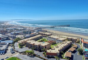 Vue aérienne d’une ville côtière avec une plage, une jetée et un ensemble de bâtiments, comprenant des parkings et des espaces verts.