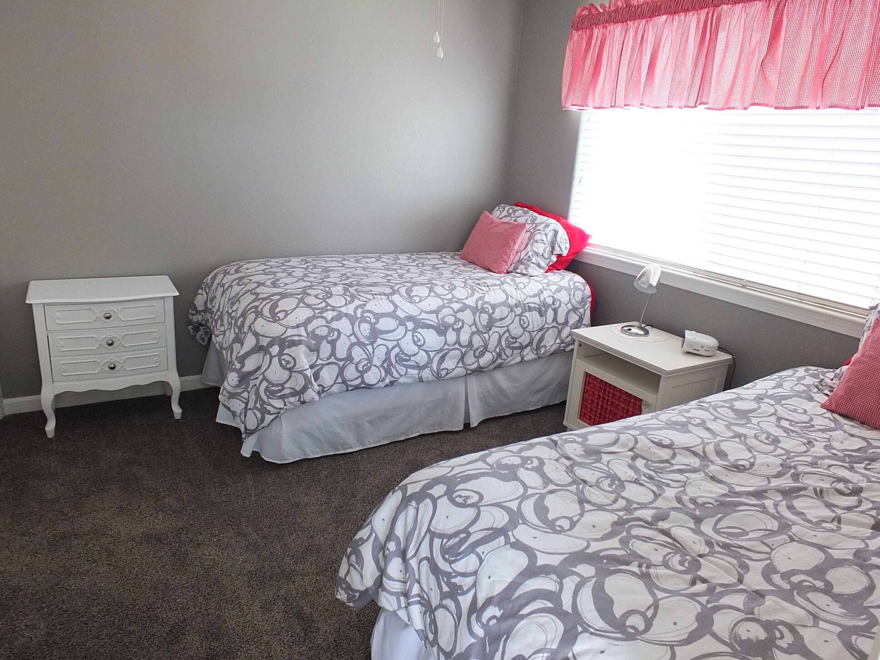 Quarto com duas camas, uma mesinha de cabeceira e uma escrivaninha com abajur.