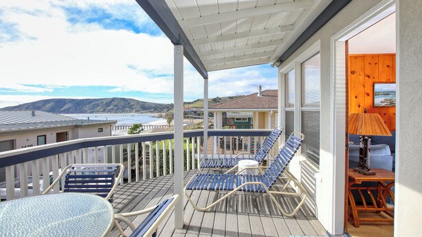 A balcony with blue and white lounge chairs, a round table, and a view of the ocean and hills. - 141 A 1st Street: 3  BR, 2  BA House in Avila Beach, Sleeps 6 (Avila Beach)