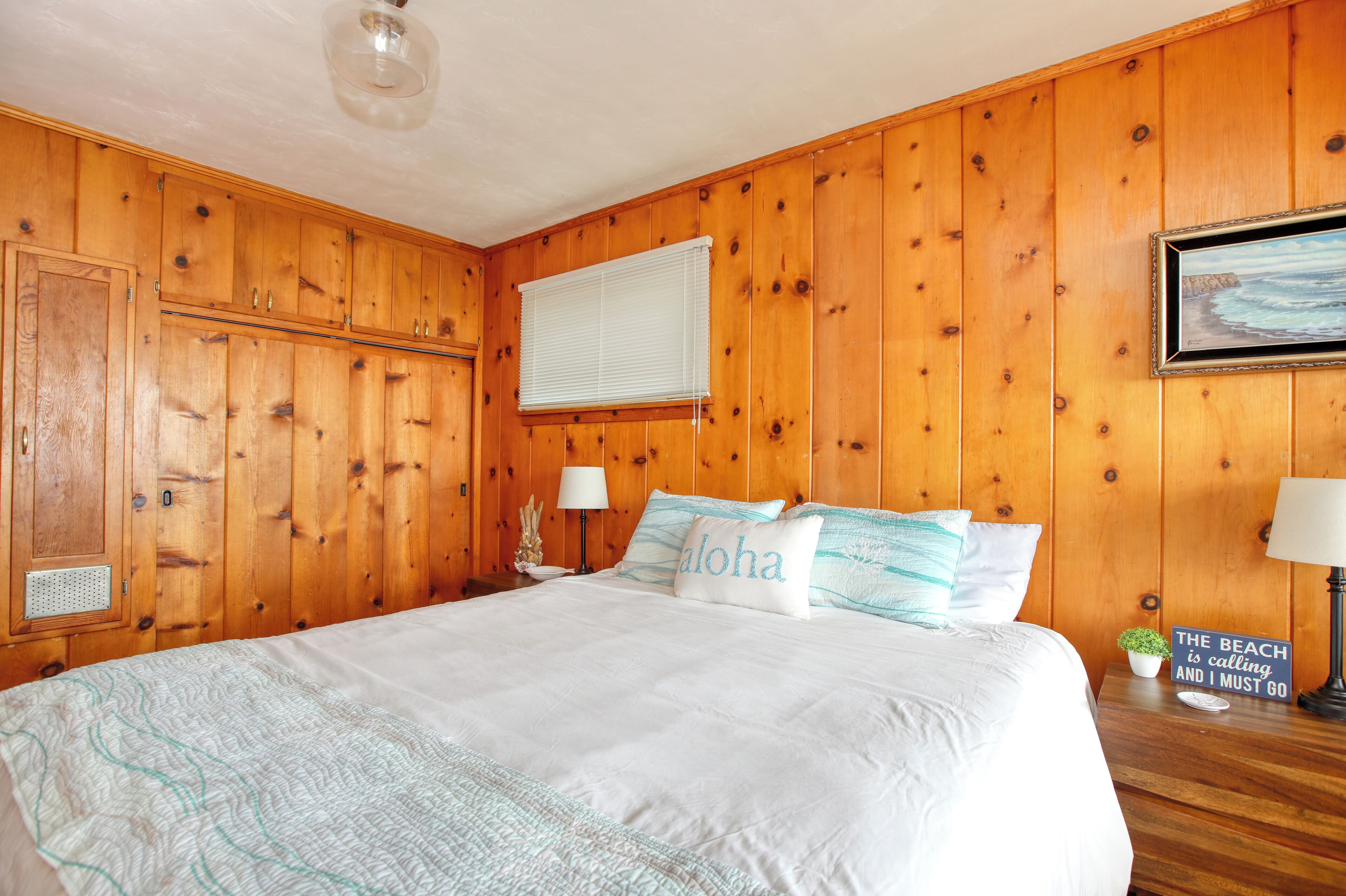 A wooden-paneled bedroom with a large bed, a nightstand with a lamp, a small plant, and a framed picture on the wall.