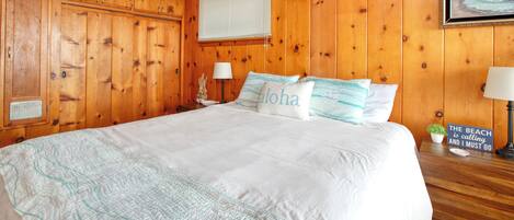 A wooden-paneled bedroom with a large bed, a nightstand with a lamp, a small plant, and a framed picture on the wall.