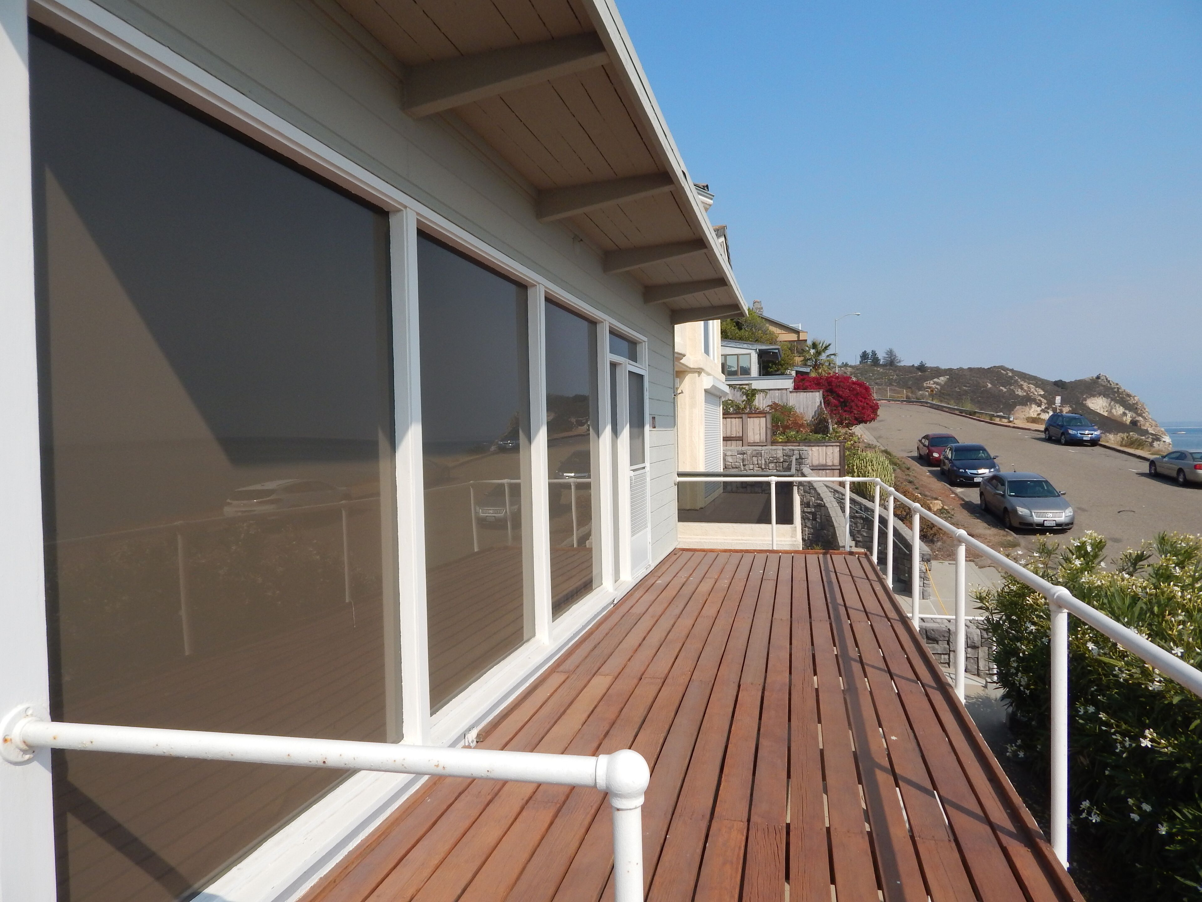 A wooden deck with a railing, overlooking a parking area and a coastal view.