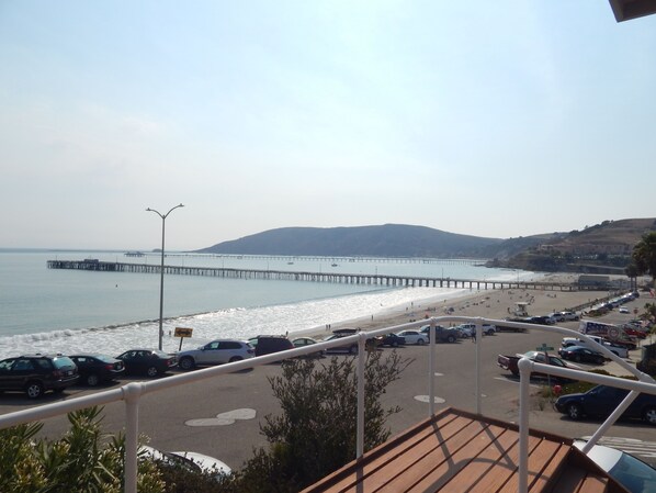 A coastal view with a long pier extending into the water, a beach with people and parked cars, and a hilly landscape in the background.