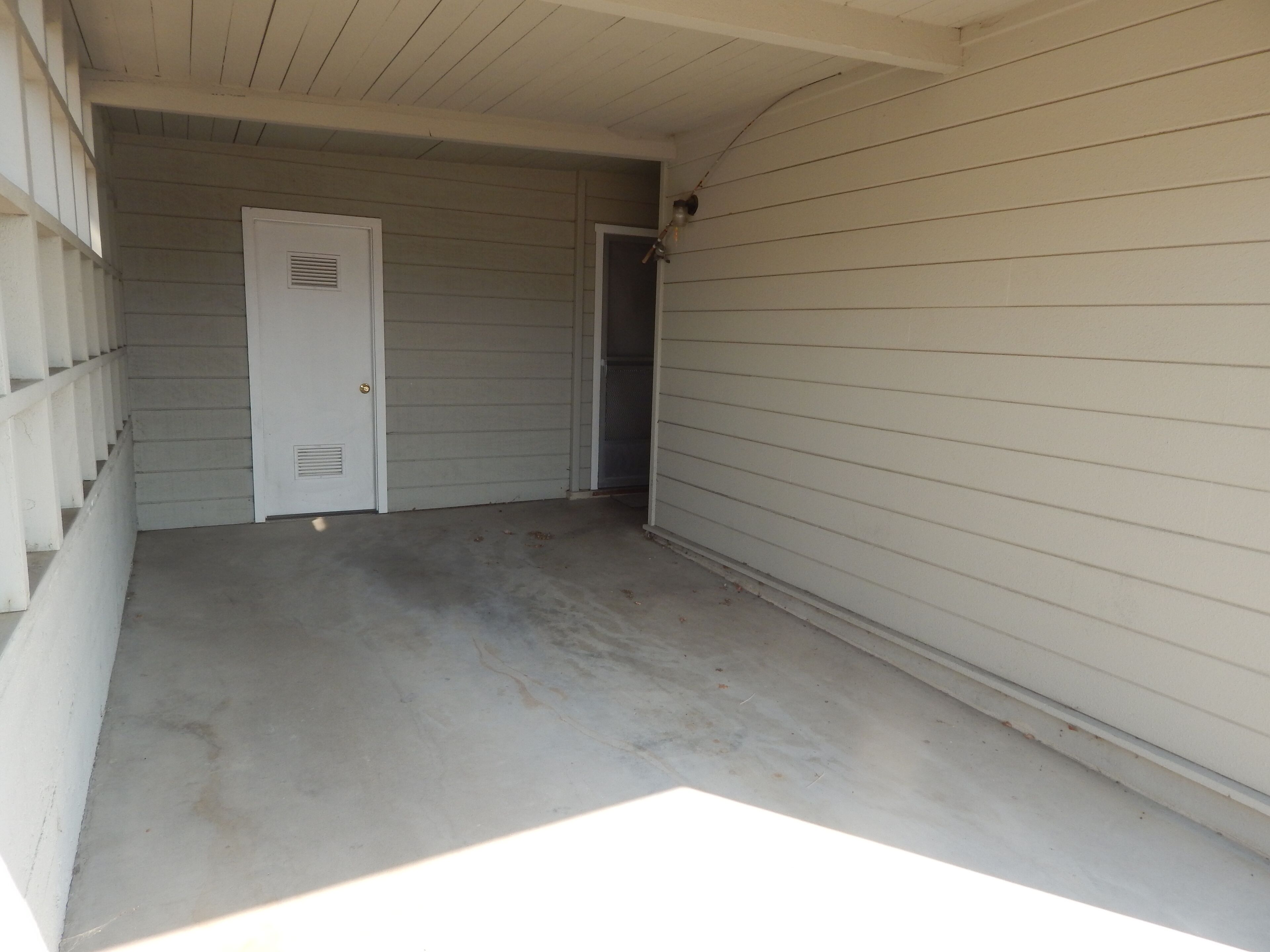 A covered outdoor area with a concrete floor, a white door, and a white exterior wall.